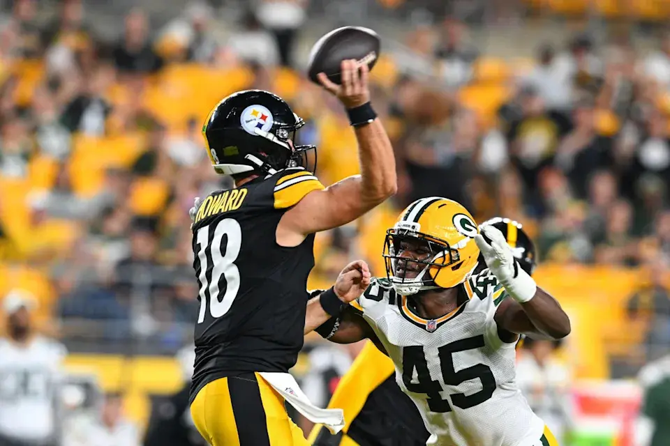Pittsburgh Steelers quarterback wearing number 18 preparing to throw a football while being pressured by a Green Bay Packers defender.