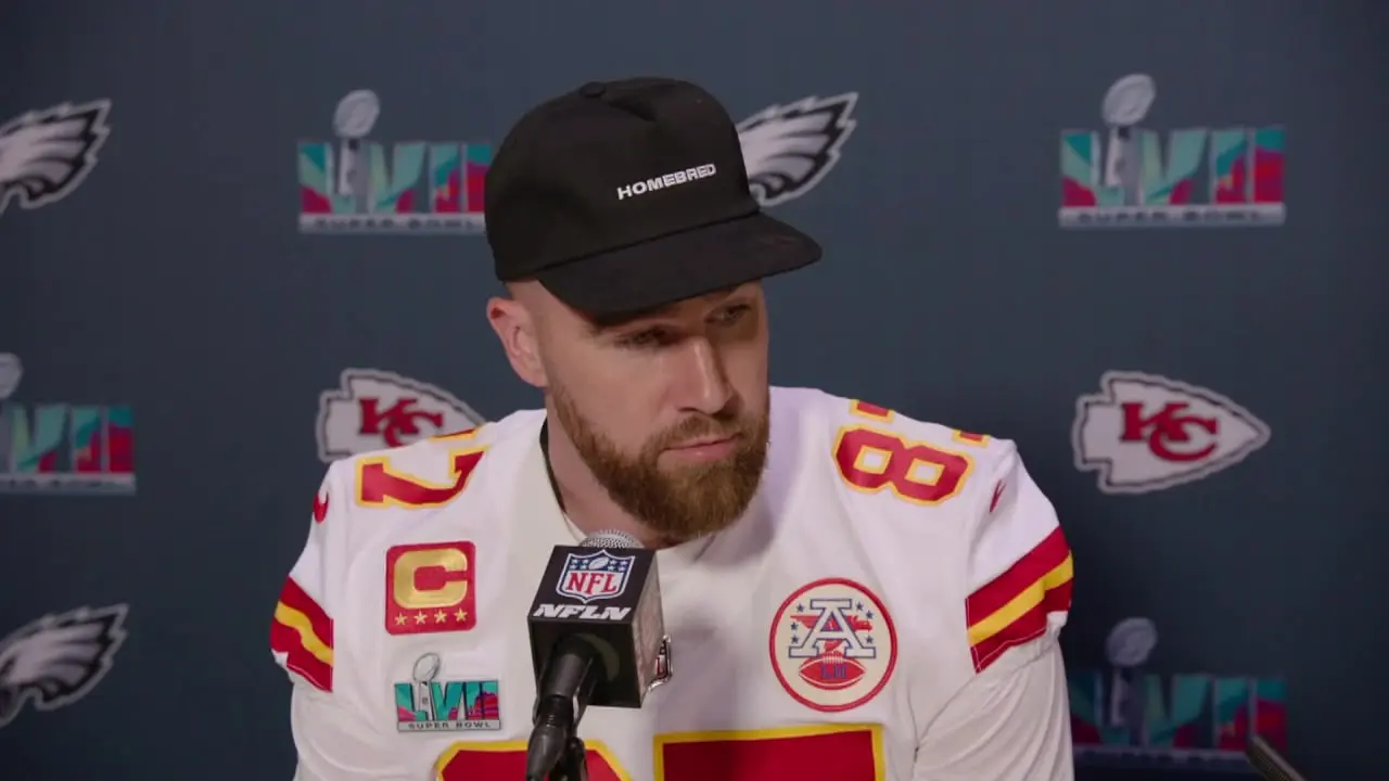 Travis Kelce wearing a Kansas City Chiefs jersey and black cap during a press conference.