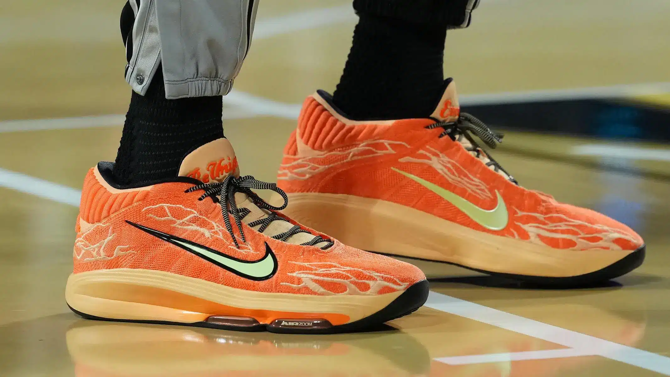 Close-up of orange Nike basketball shoes with a lime green swoosh and flame patterns on a court.